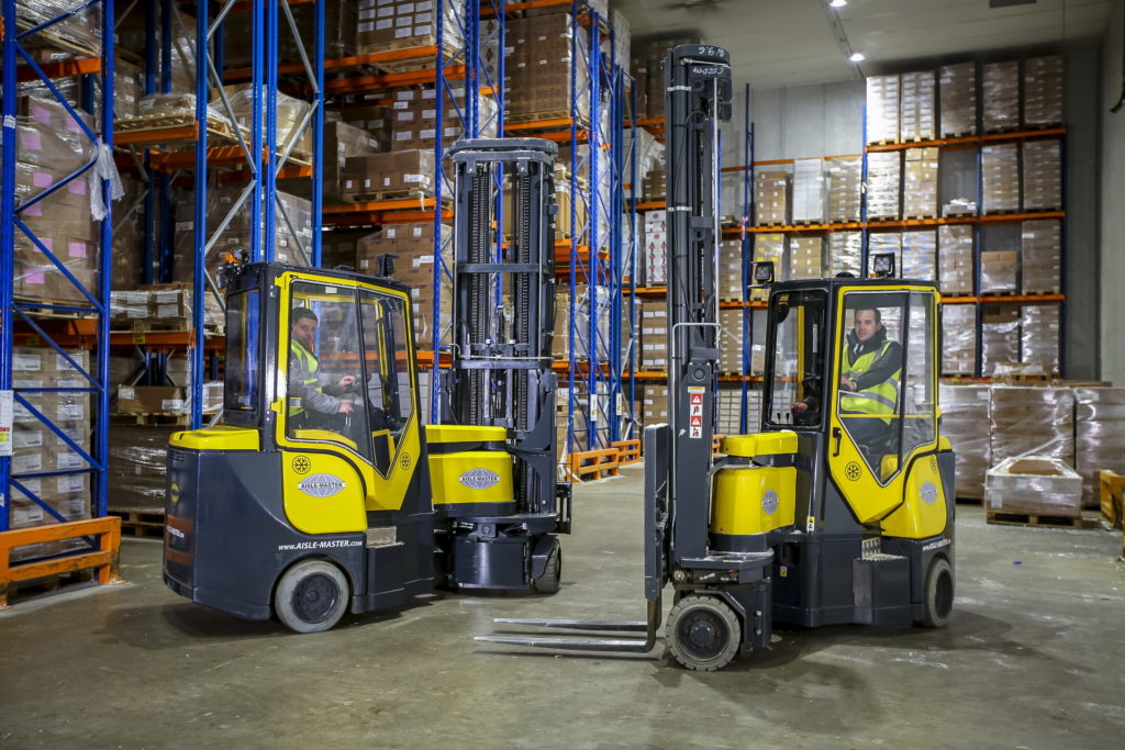 Two workers wearing high-vis operating two Aisle-Master Multi Directional Forklifts in a warehouse stacked with palettes